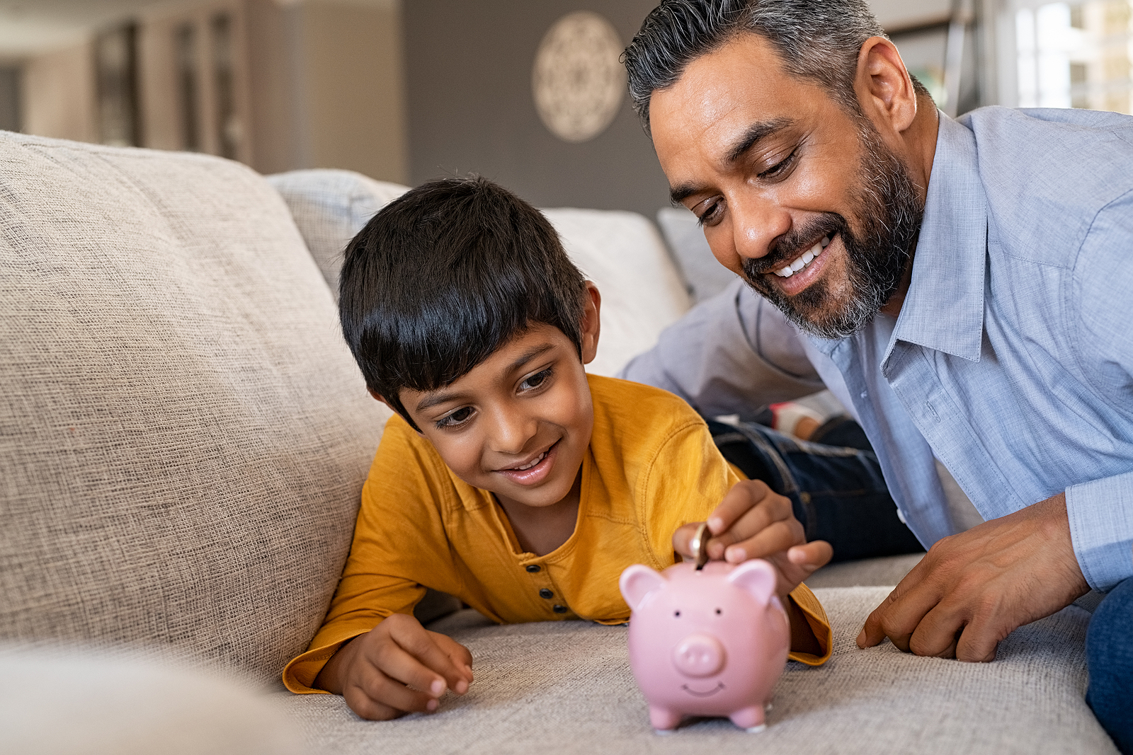Happy indian son saving money in piggy bank with father. Lovely ethnic father teaching to little boy importance of saving money for future. Smiling middle eastern kid adding coin in piggybank with dad